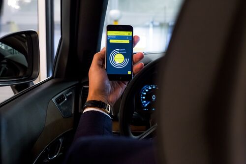A woman is sitting in a car, holding a mobile phone with an app open in front of her