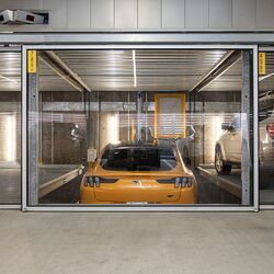 La imagen muestra un coche deportivo amarillo en un sistema de estacionamiento dentro de un moderno garaje subterráneo.